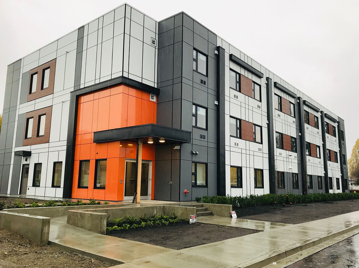 A modern, modular building on a rainy day. It's facade alternates between dark and light grey, with a splash of orange around the front door. Dark wood panels are around the second and third story windows.