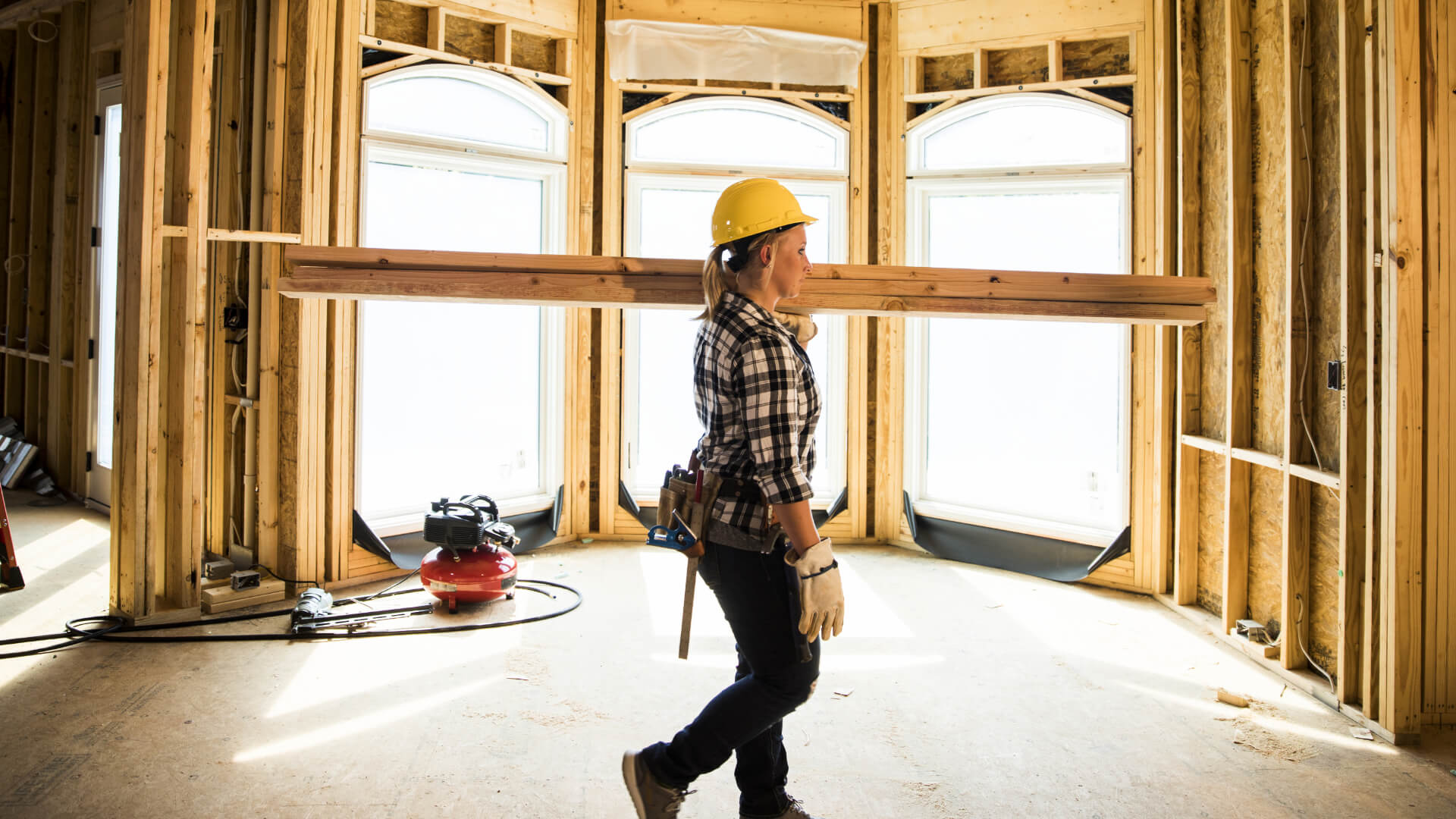 A woman in construction work carries beams of wood over her shoulder inside a newly constructed building frame.