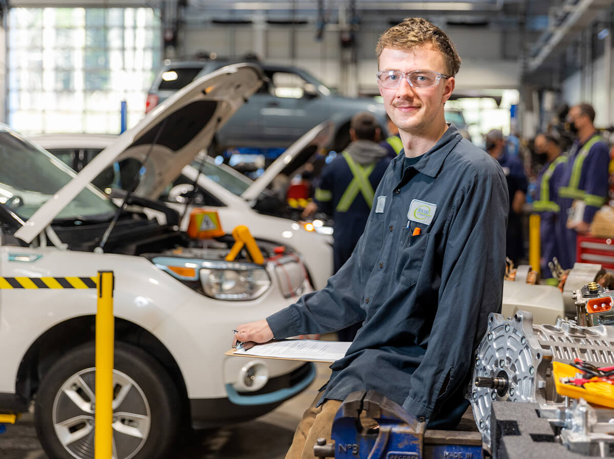 An apprentice mechanic holding a clipboard and standing beside a vehicle with an open hood while a group of students are being trained in the background.