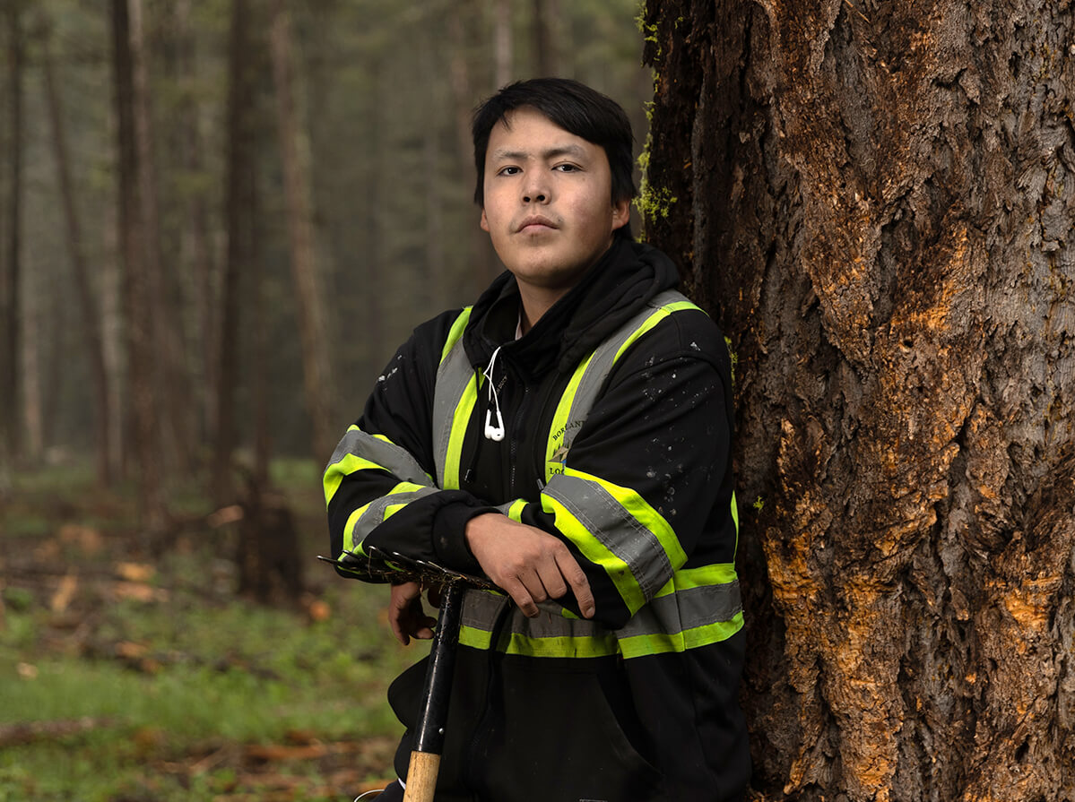 A young indigenous man wearing a reflective vest and leaning on a tool with his back to a large tree trunk.