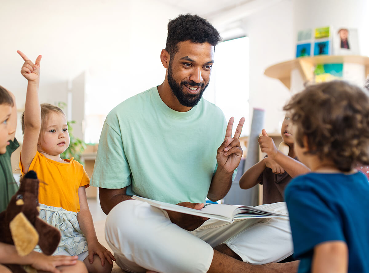 A Black man in childcare sits with children and smiles as he reads a book to them.