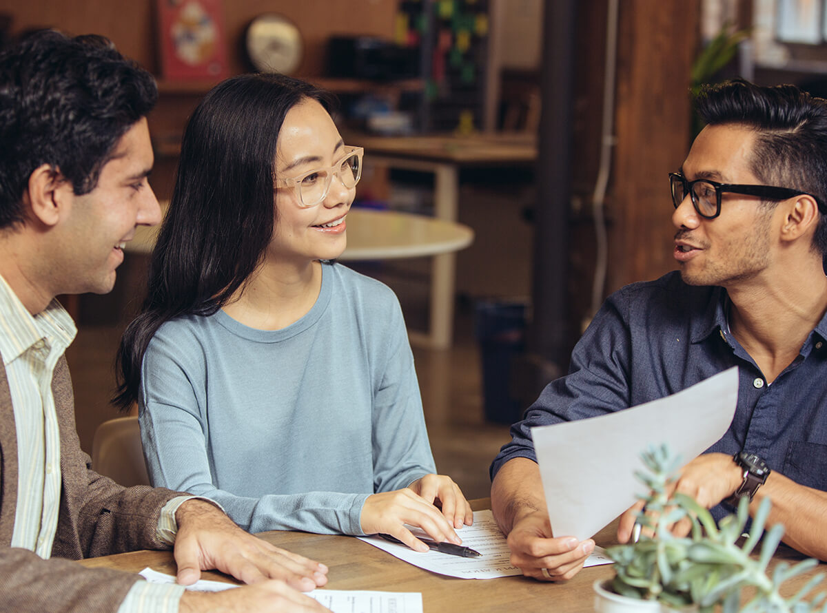 Three colleagues smiling and sitting together as they discuss printed documents.