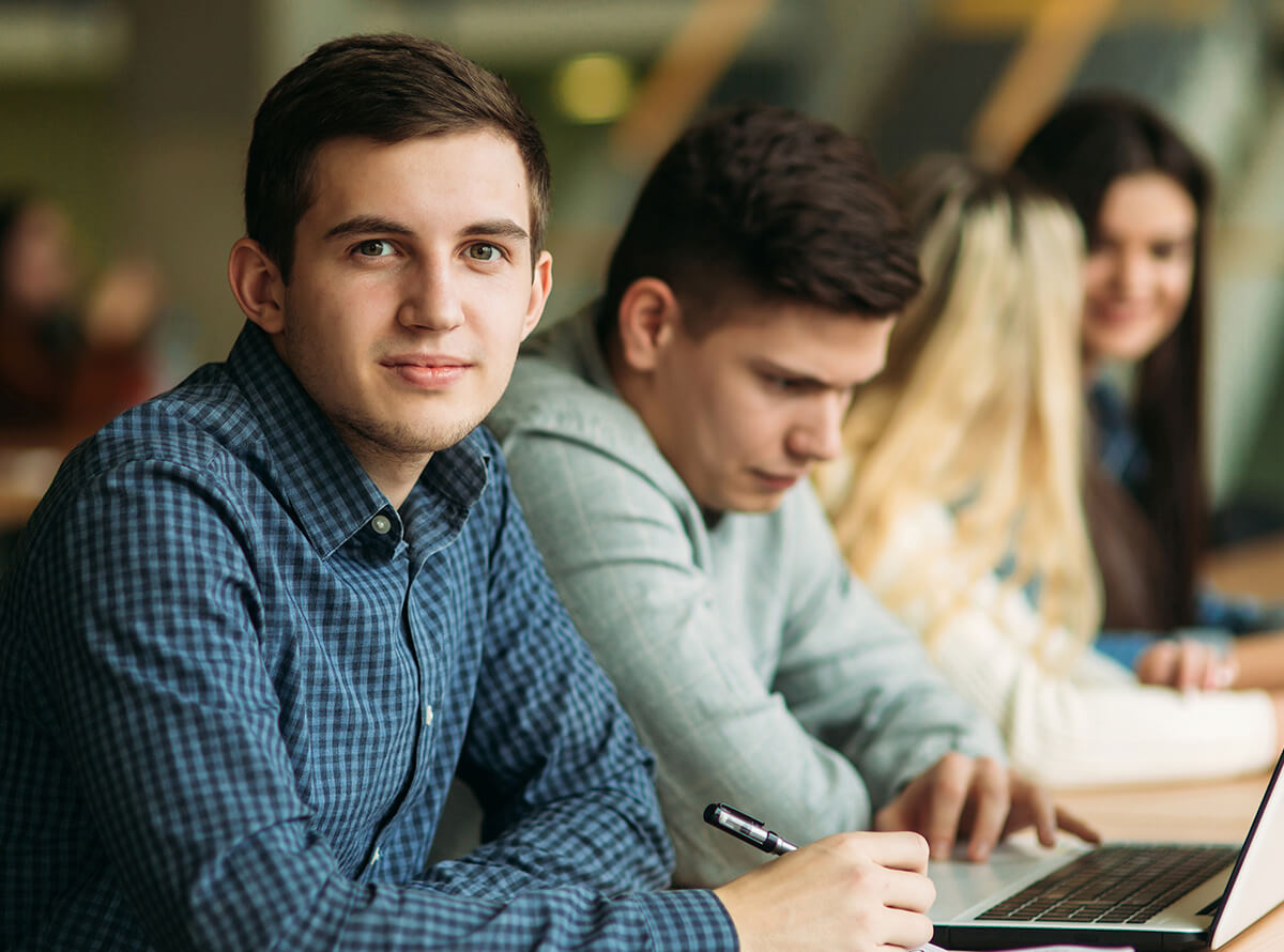 A young white man sits with a few other classmates as he looks at the camera with a pen in hand.