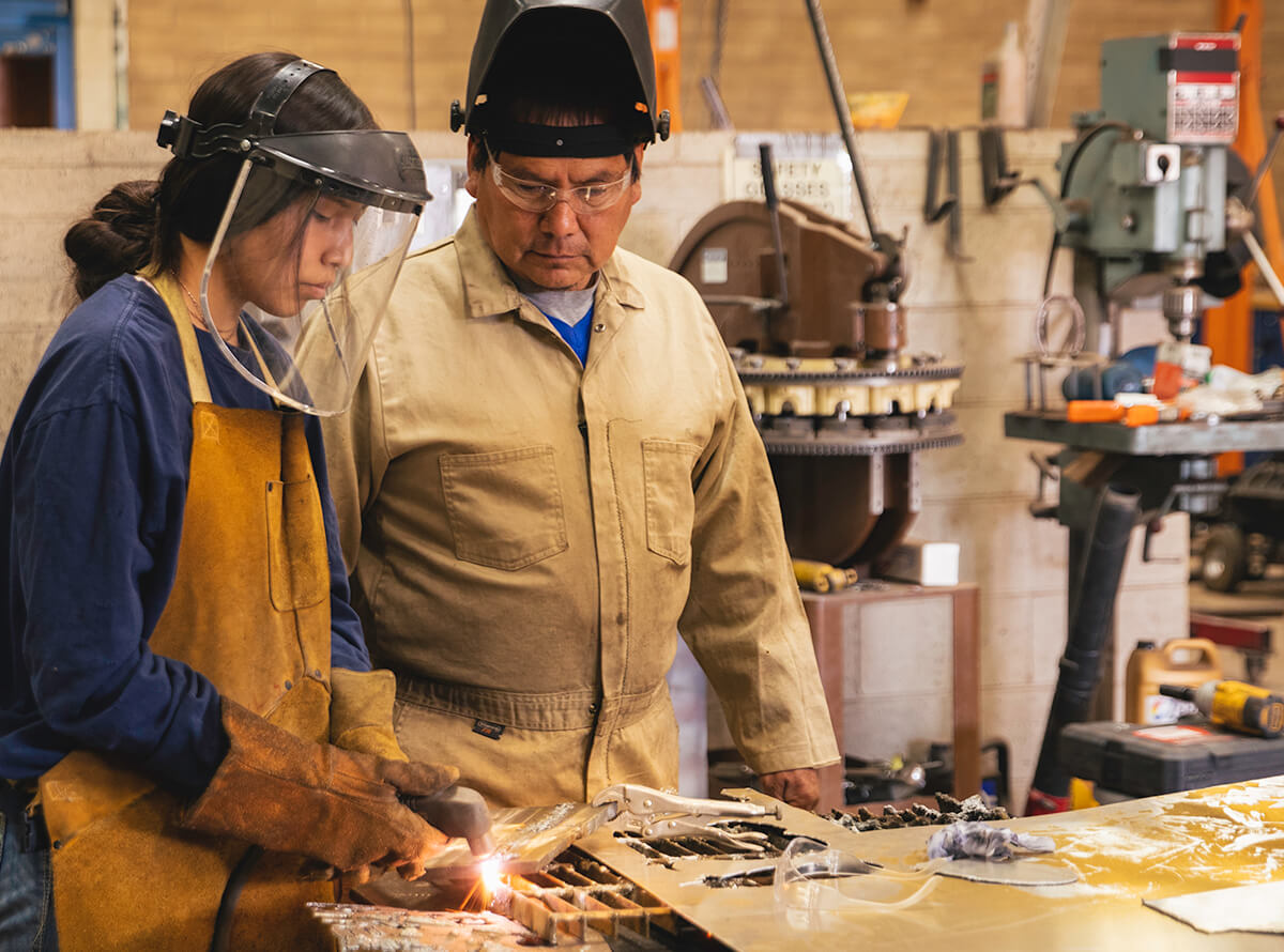 A middle-aged Indigenous man wearing goggles and coveralls watches a young Indigenous woman wearing a shield mask as she welds a piece of metal.