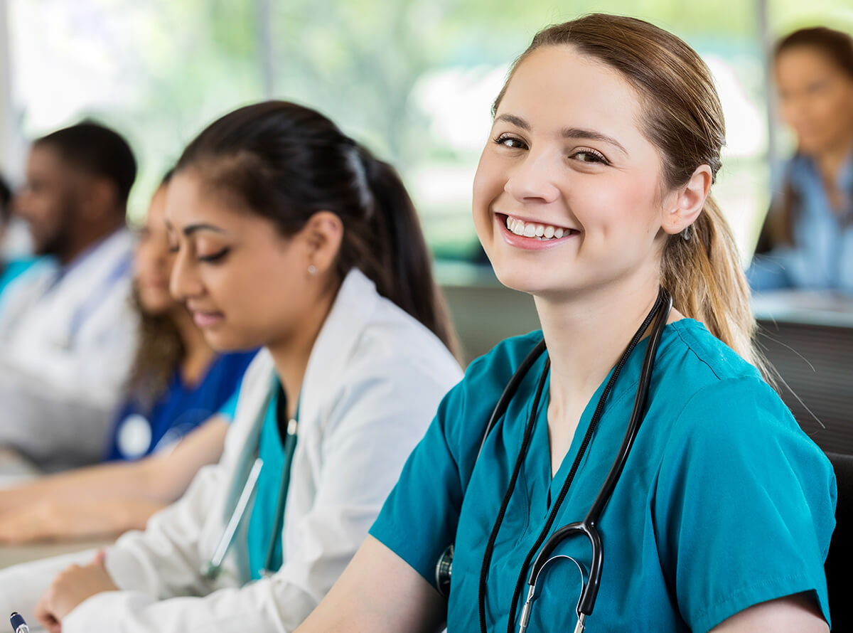 Med students wearing scrubs and stethoscopes sit in a class together. The focus is on a young white woman smiling at the camera.