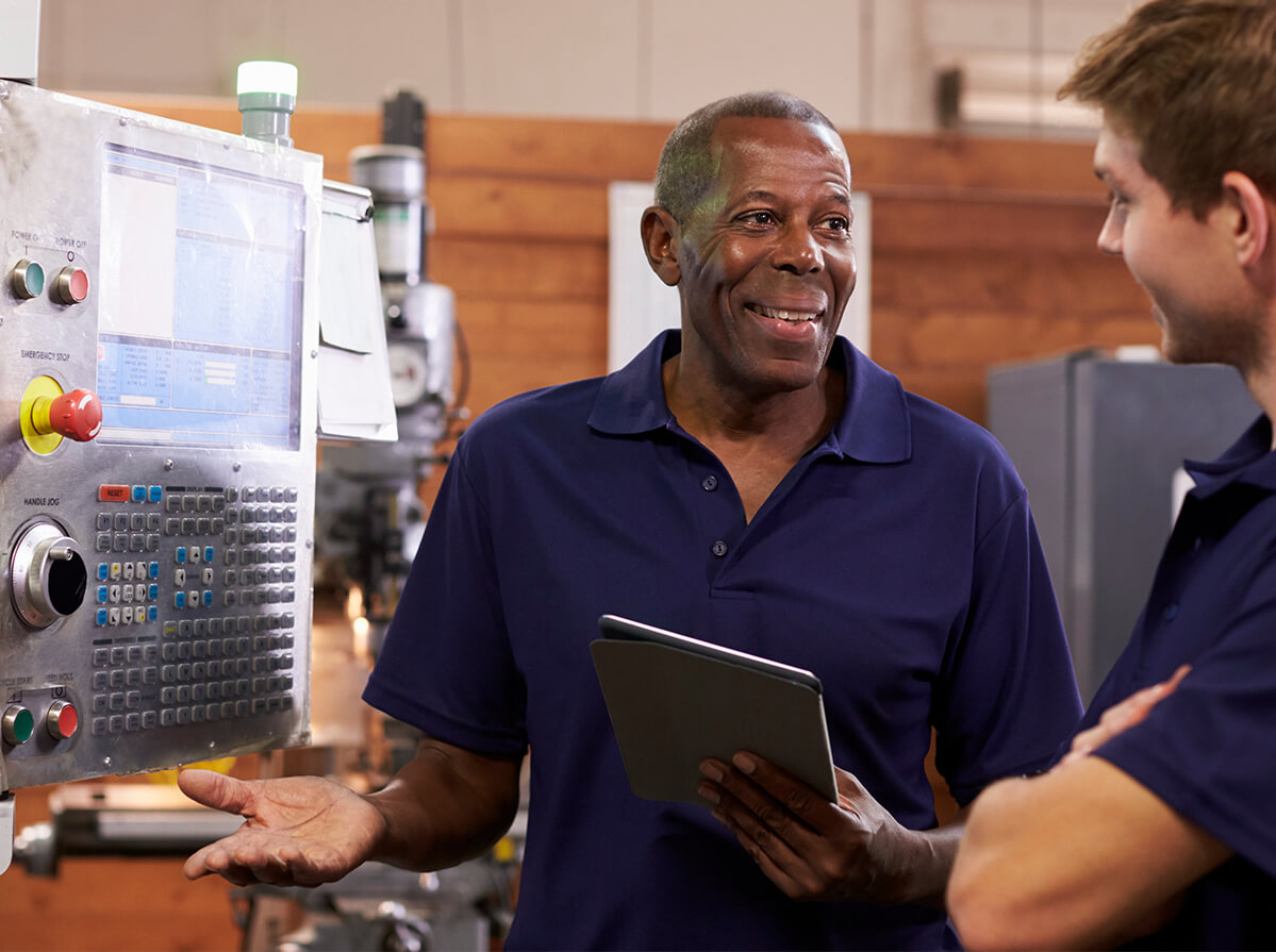 A Black man smiles at a younger colleague while they work with machinery.