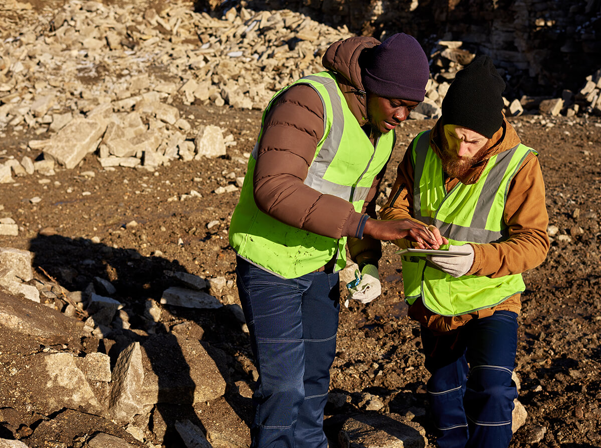 Two workers in hi-vis vests are looking at a digital document together while standing in a field of dirt and rubble on a sunny day.