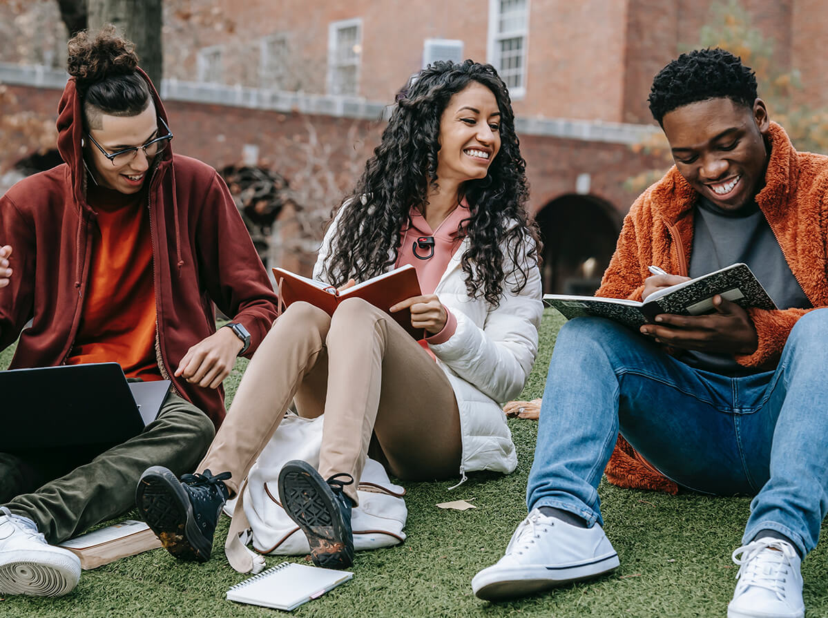 A group of young adults sit and laugh together while working on their homework and sitting on fake grass outside a post-secondary building.