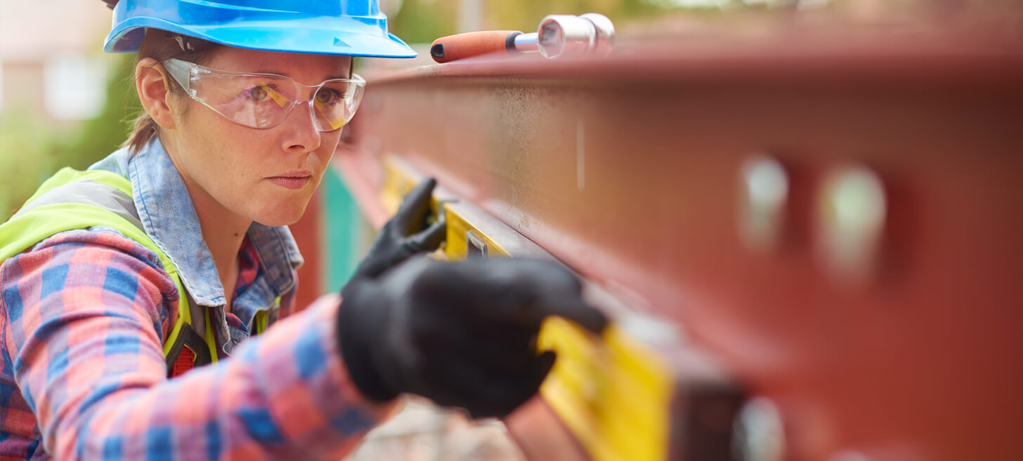 A woman wearing a hard hat, hi-vis vest and gloves uses a large level in working on a construction.