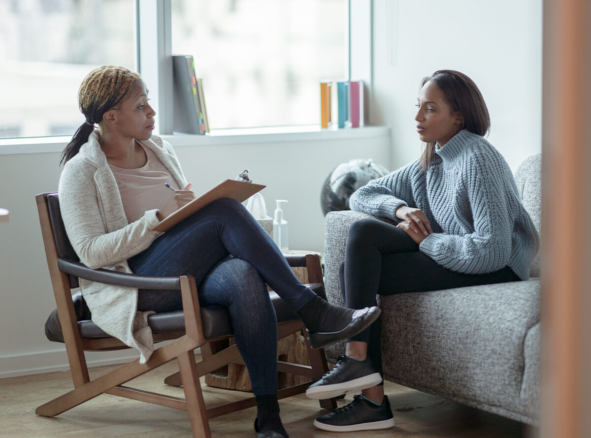 Two women sit solemnly in a mental health setting. One holds a clipboard while sitting in a chair and the other faces her from the sofa.