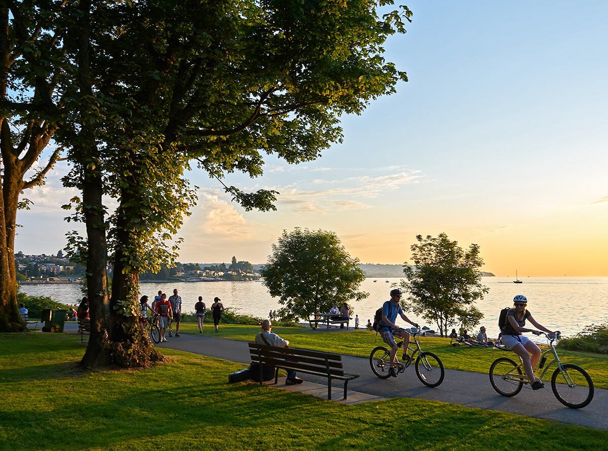 A pedestrian and cyclist paved path in a park with trees and benches overlooking the water. People are enjoying the outdoor space during sunset.
