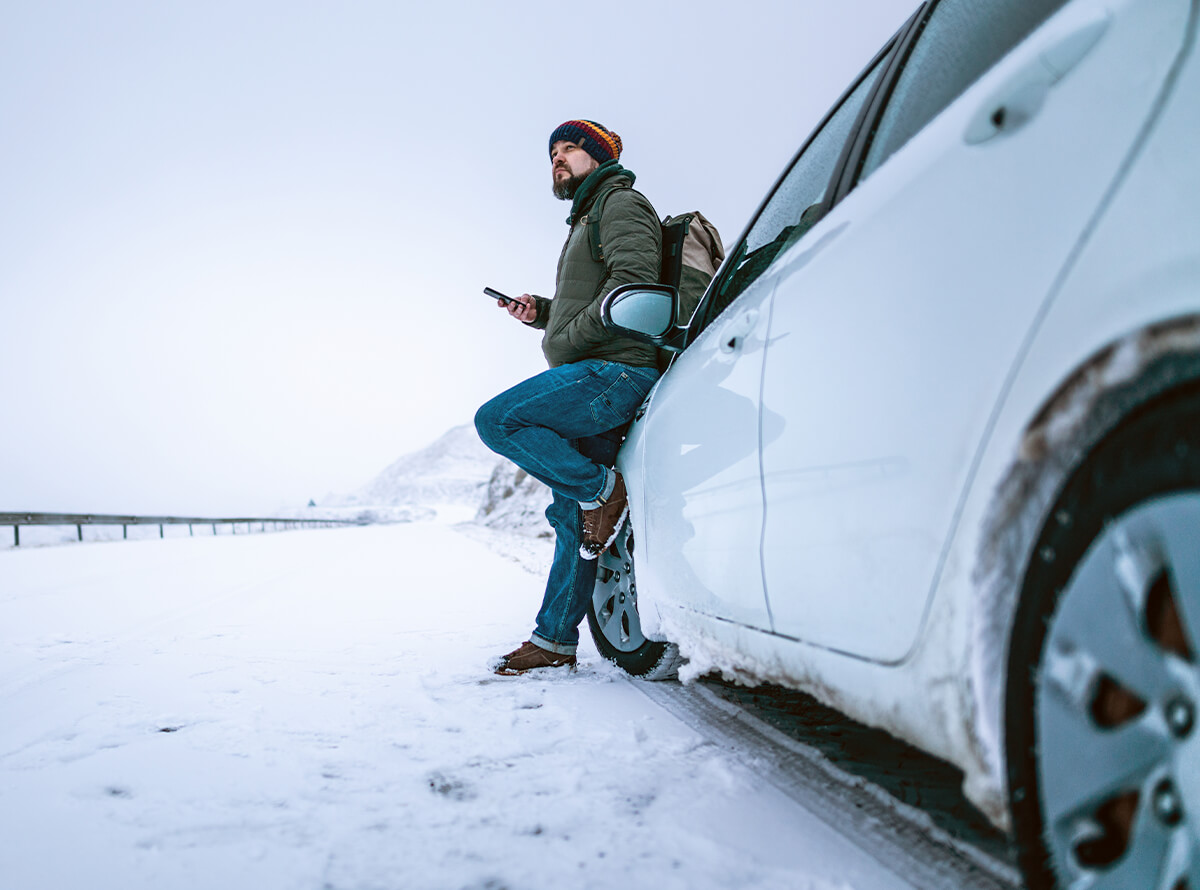 A white man leans against a vehicle with one foot up. There is snow on the ground and the sky is grey.
