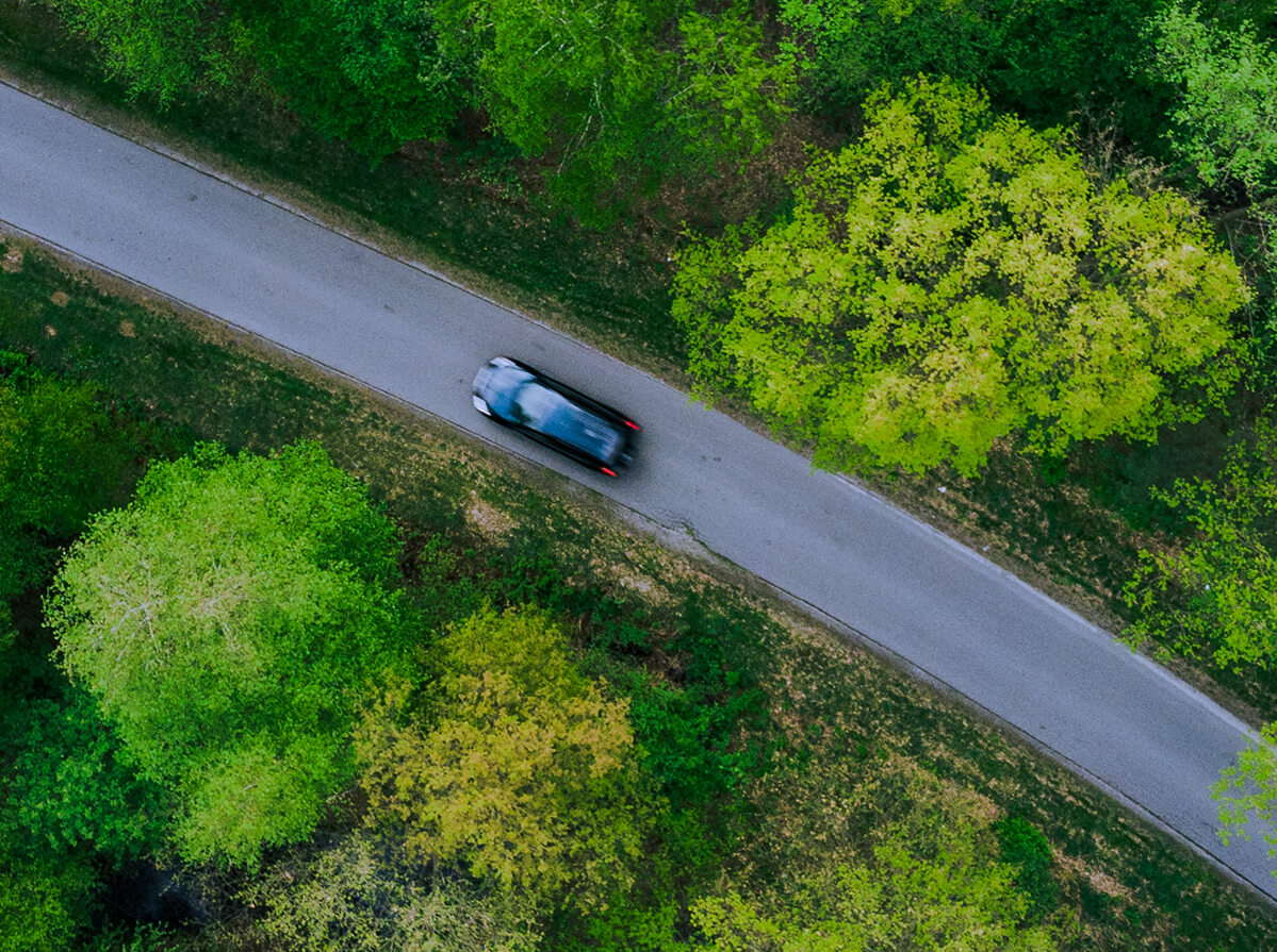 Aerial shot of a black vehicle driving on a narrow road between bright green forest.