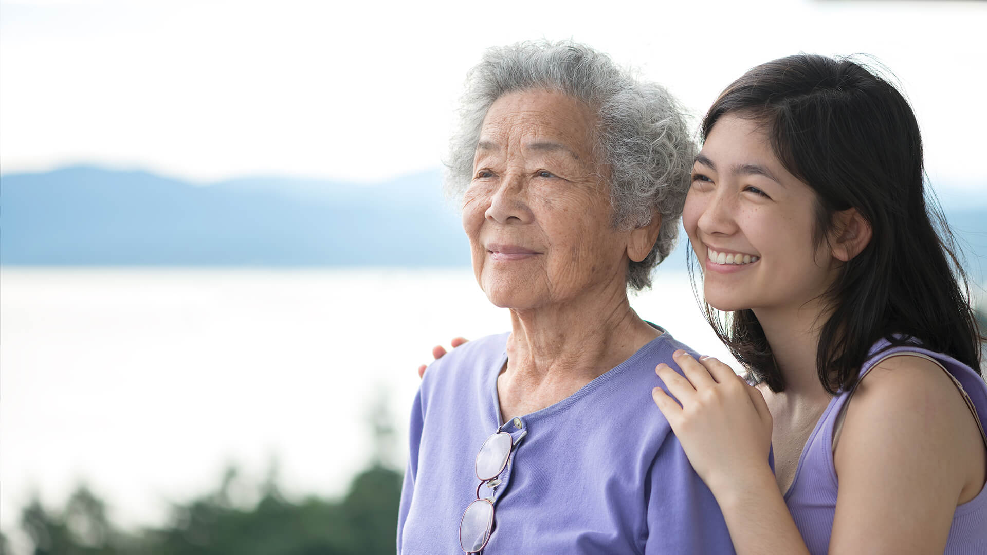 A young Asian woman and her grandmother both wear lavender shirts outside as they smile and look out into the distance together.